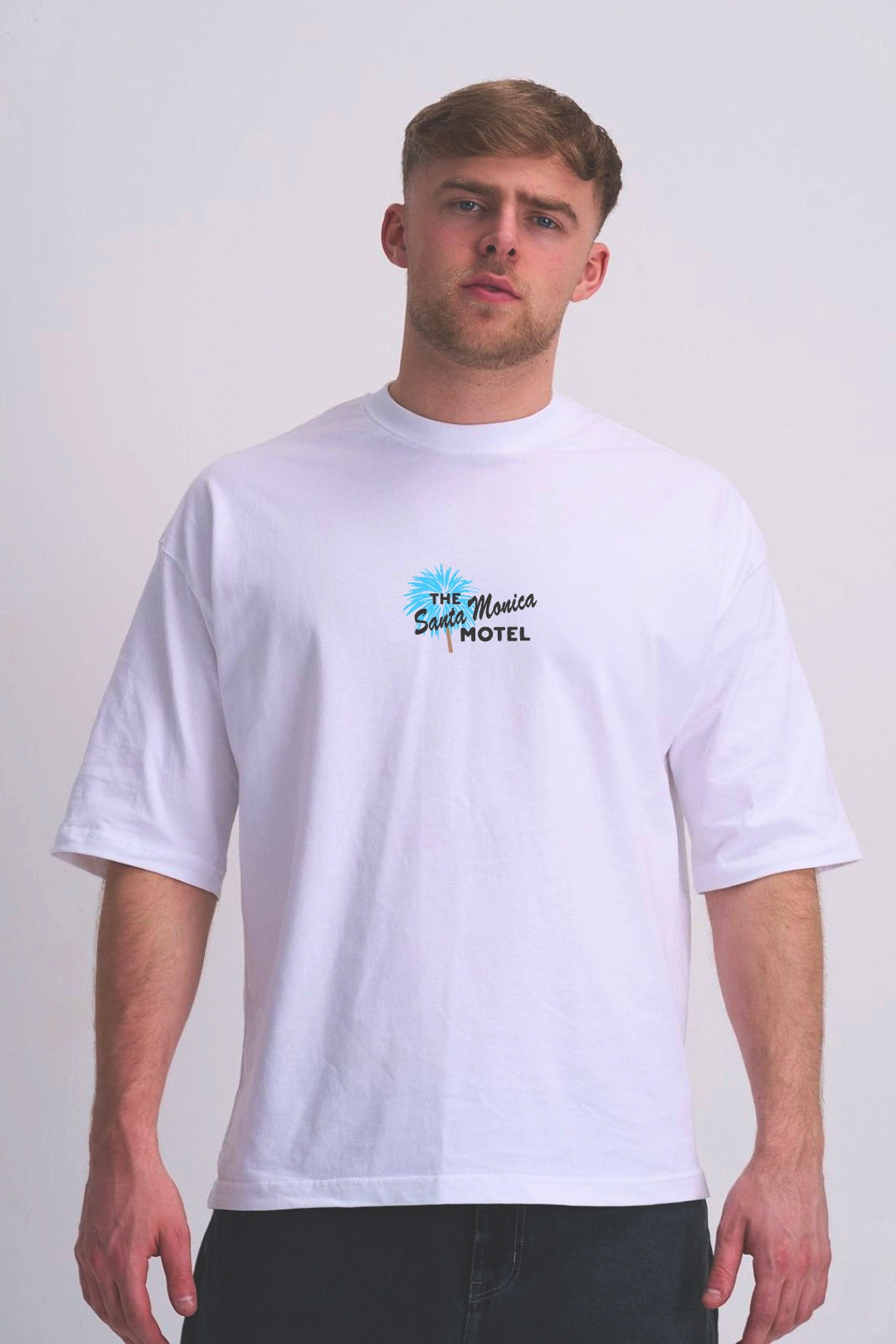 A man wearing a white short-sleeve t-shirt with a 'The Santa Maria Hotel' graphic on the chest, paired with dark grey jeans, standing against a light grey studio background.