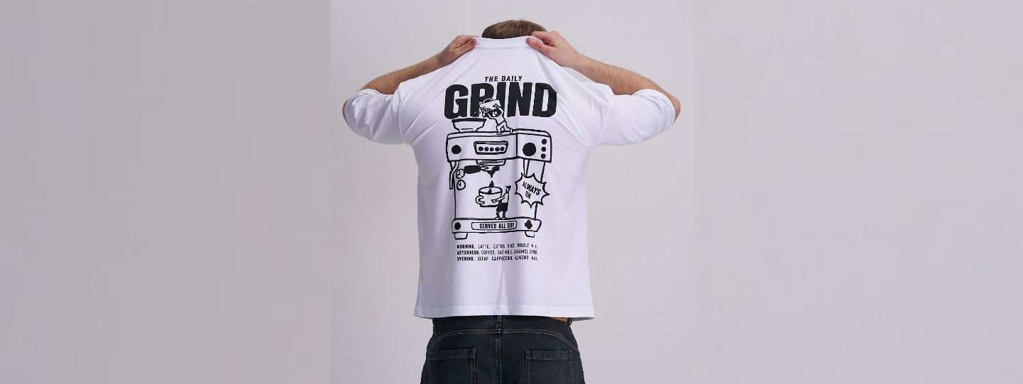 Person wearing a white t-shirt with a coffee machine graphic design and text on a plain background