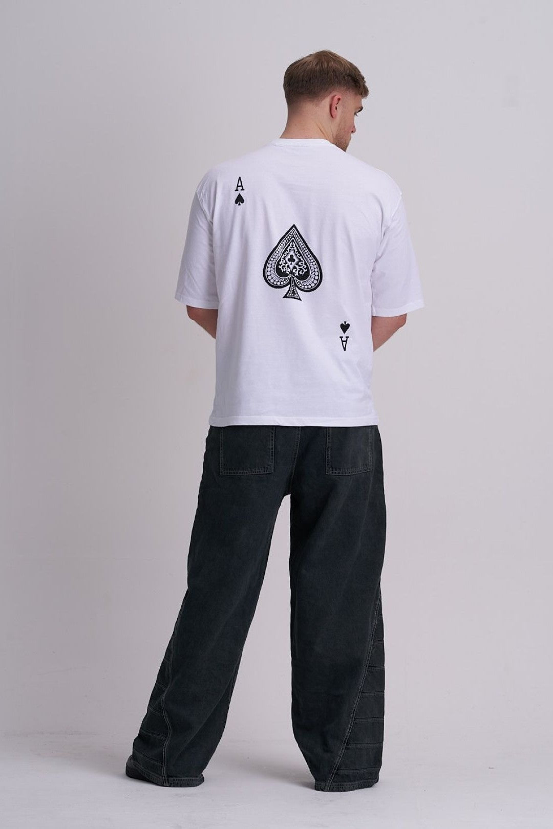 Man wearing a white t-shirt with a black spade symbol on the back, standing against a plain background.