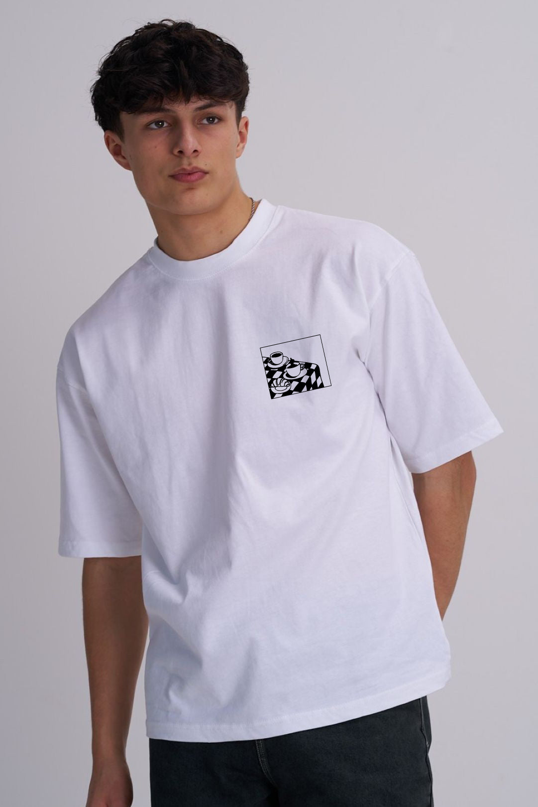 A young man wearing a white short-sleeve crewneck t-shirt with a small black and white coffee cup graphic print on the left chest, paired with dark jeans, against a plain light grey studio background.