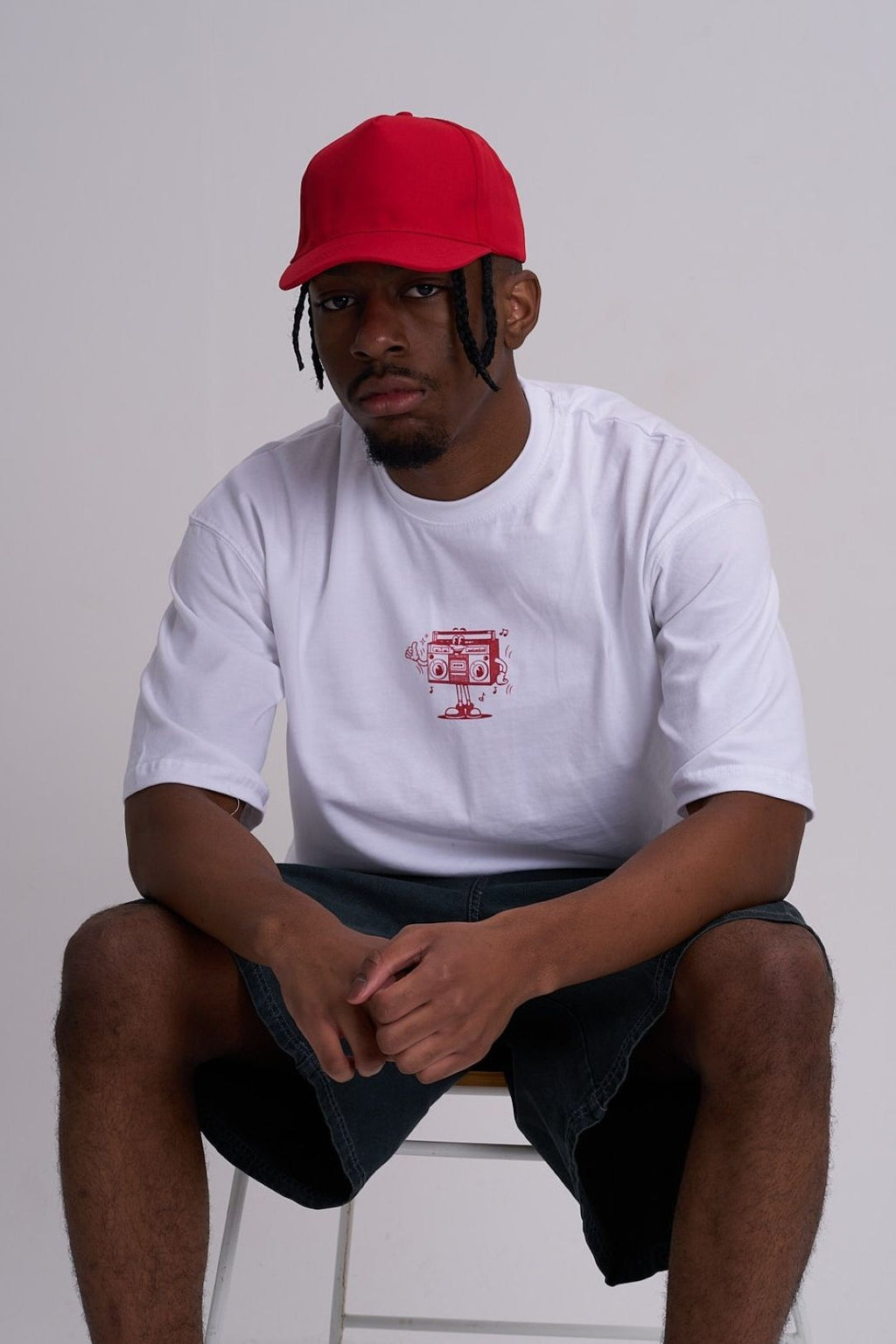 Male wearing a white t-shirt with a red retro music logo and a red cap, sitting on a white stool against a plain background.