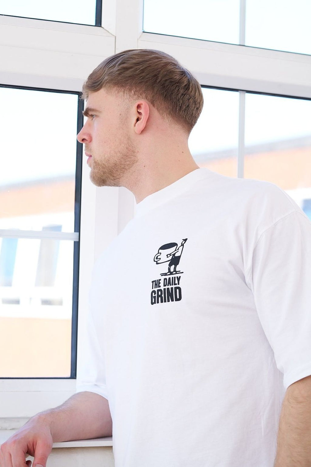 Man wearing a white t-shirt with a coffee graphic and text, sitting by a window.