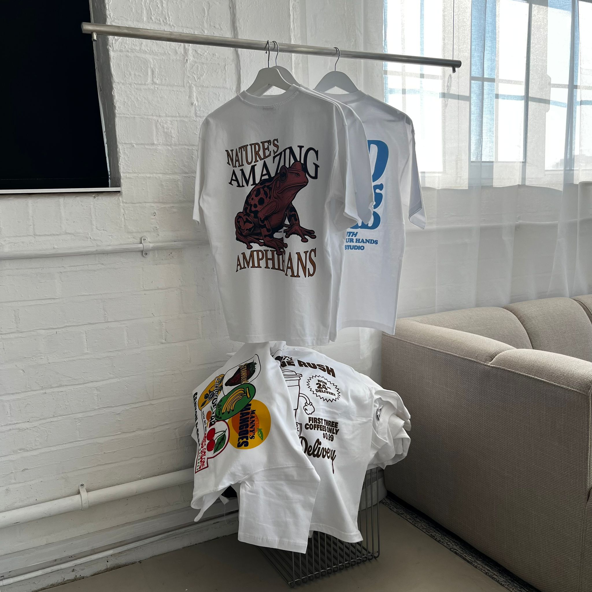 White t-shirts with various designs hanging on a rack against a white brick wall.